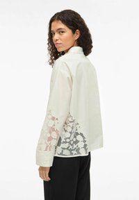 Balta kokvilnas krekls ar izšūtu ziedu mežģīnēm uz apakšmalas, garām piedurknēm un brīvi piegriezumu. Iespējas ir apkakle un pogāmas aproces.