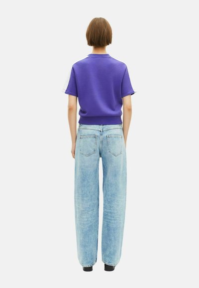 Osoba widziana z tyłu, ubrana w fioletową bluzkę z krótkim rękawem i jasnoniebieskie dżinsy w luźnym kroju, na białym tle.