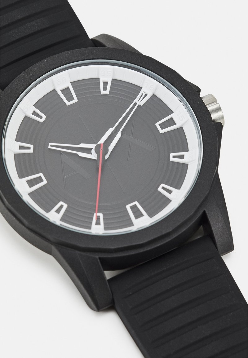 Reloj negro con correa de goma, esfera redonda negra, marcadores de hora blancos, un segundero rojo y una corona plateada. Presenta una correa texturizada.
