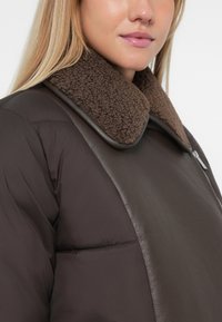 Jachetă puffer maro cu guler din blană maro și detalii din piele. Material moale, texturat, cu un finisaj neted, design călduros.