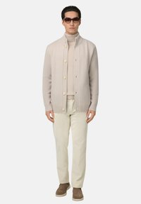 Ljust beige cardigan med ribbade mudd och fåll, med stora knappar. Bärs över en matchande polotröja och tillsammans med kritvita byxor.