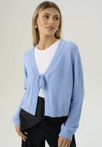 Donna che indossa orecchini a cerchio d'oro, un cardigan azzurro chiaro annodato davanti su una maglietta bianca e pantaloni neri, posa davanti a uno sfondo neutro.