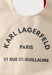 Bežová taška s čiernym textom "Karl Lagerfeld Paris 21 Rue St-Guillaume" a viditeľným červeným lemovaním textilnej rukoväte.