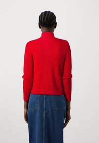 Pull rouge en maille côtelée avec col montant et manches longues, associé à une jupe en jean avec deux poches à l'arrière.