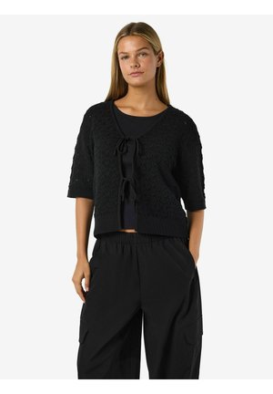 Mujer con un cárdigan de punto negro de manga corta con cierres de lazo sobre una camiseta negra y pantalones cargo negros holgados, de pie con una mano en el bolsillo.