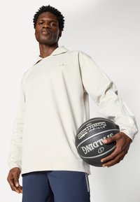 Polo de manga longa creme com colarinho plano, apresentando um logótipo discreto e três riscas brancas em cada manga. Segura uma bola de basquetebol preta.