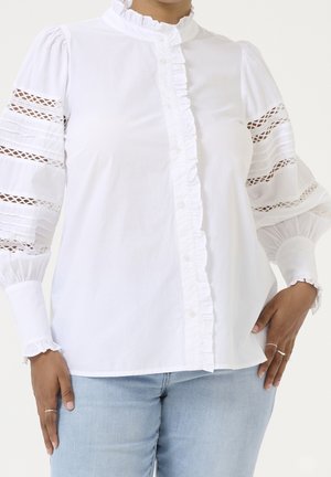 Witte blouse met gerimpeld voorpand en poflange mouwen met kanten inzet, gedragen met lichtblauwe spijkerbroek, handen op de heupen.
