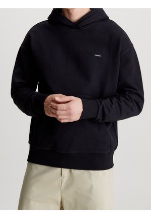 Mann trägt schwarzen Calvin Klein Hoodie mit Kängurutasche und beiger Hose, steht mit verschränkten Händen vorne.