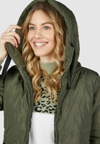 Veste matelassée verte à capuche avec fermeture éclair, présentant une texture lisse. Portée sur un pull en maille à motifs avec un design léopard.