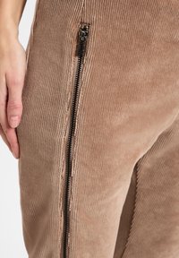 Bruine ribfluweel broeken hebben een zijrits met zwarte hardware, tonen een verticale ribstructuur en een aansluitend ontwerp langs het been.