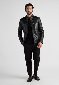 Zwarte leren jas met een getailleerd ontwerp, ritssluiting aan de voorkant, hoge kraag en twee borstzakken. Gemaakt over een zwart shirt en gecombineerd met zwarte broek.