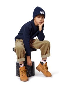 Pull en maille bleu marine, pantalon en velours côtelé beige et bottes beiges à lacets. Les accessoires comprennent un bonnet sombre avec un logo. Assis sur un tabouret tacheté.