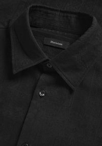 Chemise noire à col, avec un subtil motif texturé, comportant une étiquette portant le nom "Matinique" et des boutons noirs à l'avant.