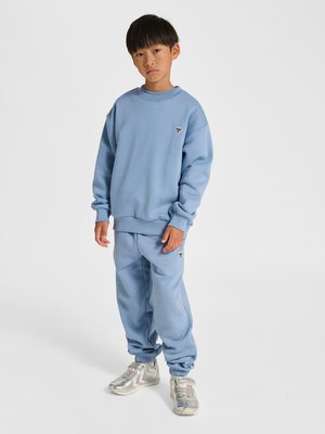 Jonge jongen staat, draagt een lichtblauwe trui en bijpassende joggingbroek met kleine zwart-witte logo's, en zilveren sneakers.