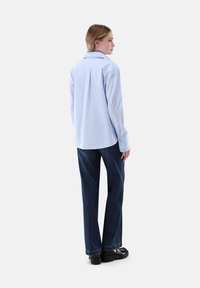 Donna vista di lato che indossa una camicia a maniche lunghe azzurra, jeans blu scuro e scarpe nere su uno sfondo semplice.