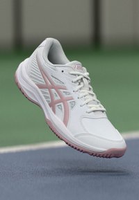 Hvit og rosa Asics treningssko i luften over et blått og grønt idrettsbanedekke.