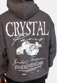 Szara, przyduża bluza z kapturem z dużym białym napisem na plecach: "CRYSTAL Paryż", grafiką diamentów oraz napisem "OSTROŻNIE" w mieszanych czcionkach.