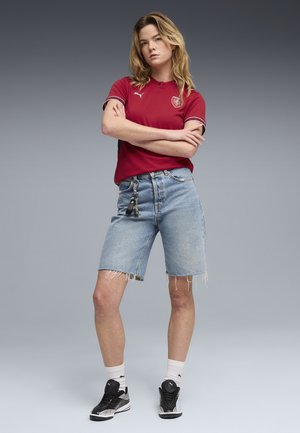 Jeune femme bras croisés portant un maillot de sport rouge, un short en jean avec porte-clés, des chaussettes blanches et des baskets Puma noires, sur un fond gris.
