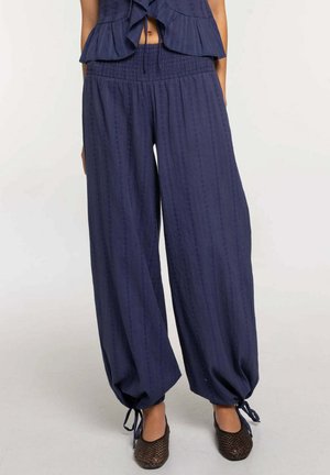 Mujer con pantalones anchos azul oscuro con ataduras en los tobillos y zapatos negros tejidos tipo slip-on sobre un fondo liso claro.