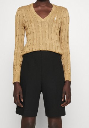 Personne portant un pull beige en maille torsadée col V et un short noir taille haute longueur genou sur un fond uni.