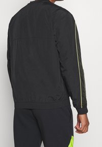 Svart bomberjacka med en slät textur, har neongula sidostraps, elastiska muddars och en ribbad midja. Bakvy visas.