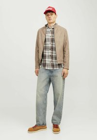 Een tan suède jasje over een geruit shirt, lichtblauwe baggy jeans en bruine laarzen met rode veters; een rode pet met witte tekst.