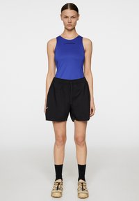 Blå ärmlös topp med logotyp, matchad med svarta shorts med elastisk midja. Modellen bär beige skor med färgglada detaljer.