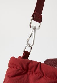 Primo piano di una borsa di tessuto rosso, con un moschettone in metallo argento e una tracolla regolabile. La texture appare spiegazzata con cuciture visibili.