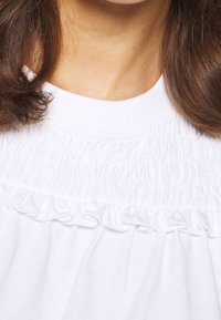 Vit topp med en samlad smockdesign och volanger vid halsen. Mjuk textur i en lös passform, perfekt för avslappnad användning.