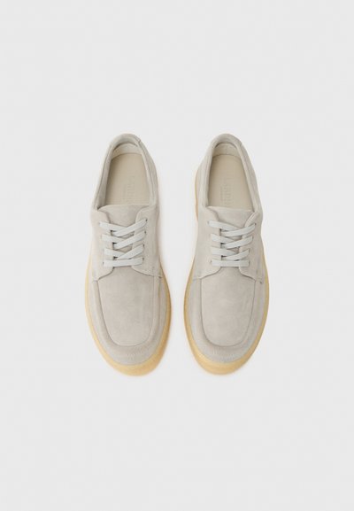 Adidași gri din suede cu vârf rotund, șireturi gri deschis și o talpă din cauciuc bej masivă. Prezintă un design minimalist fără logo-uri vizibile.