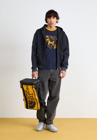 Črna jakna na zip nad temnomodrim majicama s potiskom, temne jeans hlače in srebrni športni copati. Oseba drži črno in rumeno torbo z logotipom.