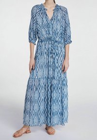 Maxime-kleid in Blau mit einem Ikat-Muster, leichtem Stoff, Bindeband in der Taille und Dreiviertelärmeln. Einfache Sandalen runden den Look ab.
