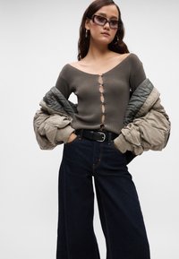 Νεαρή γυναίκα φορώντας μια oversized μπεζ ζακέτα κρεμασμένη στους ώμους, καφέ ριγωτό τοπ με κουμπιά, σκούρα φαρδιά τζιν παντελόνια, μαύρη ζώνη και ορθογώνια γυαλιά ηλίου.