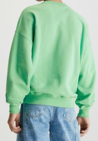 Mátově zelená mikina s kulatým výstřihem, oversized siluetou a žebrovanými manžetami. V kombinaci s světle modrými džínsovými šortkami.