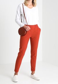 Woman wearing white long-sleeve shirt, burnt orange pants, white slip-on shoes, and a round brown crossbody bag.