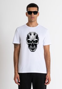 Camiseta blanca de algodón con un gráfico de calavera en negro y plata con ojos verdes, combinada con gafas de sol negras. Diseño sencillo y casual.