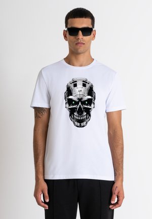 Camiseta blanca de algodón con un gráfico de calavera en negro y plata con ojos verdes, combinada con gafas de sol negras. Diseño sencillo y casual.