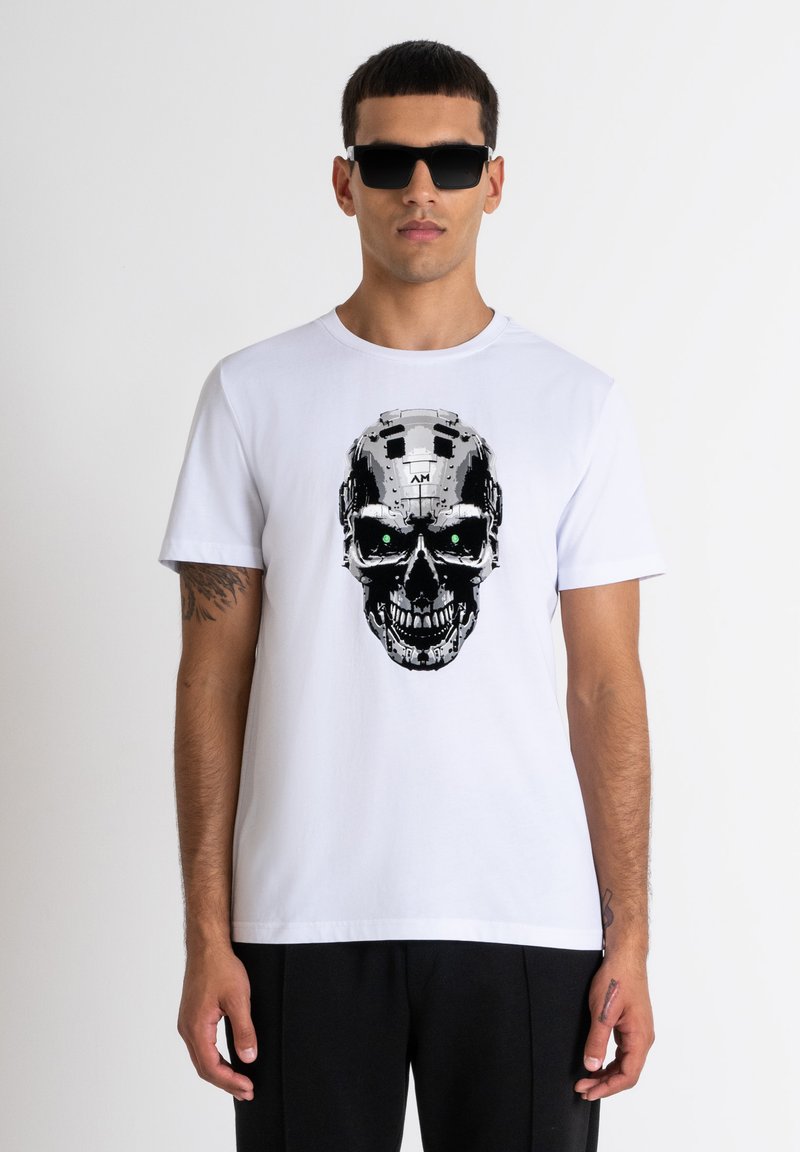 Camiseta blanca de algodón con un gráfico de calavera en negro y plata con ojos verdes, combinada con gafas de sol negras. Diseño sencillo y casual.