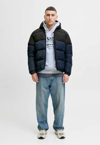 Pufferjakke i sort og navy med quiltede sektioner og hætte, båret over en grå hættetrøje. Kombineret med løse lyse jeans og sneakers.