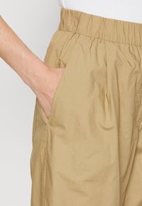 Mano infilata nella tasca laterale di pantaloni casual beige con vita elastica, indossati con una maglietta bianca visibile in vita.