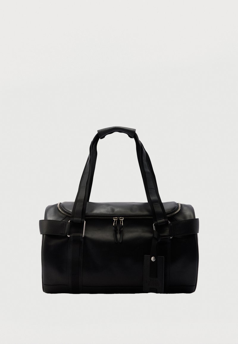Μαύρη δερμάτινη τσάντα duffel με διπλές λαβές, κλείσιμο με φερμουάρ στην κορυφή, πλαϊνά λουριά και προσαρτημένη ορθογώνια ετικέτα αποσκευών σε απλό φόντο.