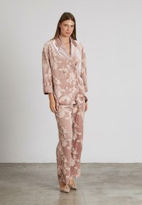 Una donna indossa un blazer floral in velluto rosa soft e pantaloni a gamba larga abbinati, completando il look con dei tacchi a punta, il tutto in un ambiente neutro.