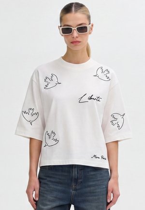 Femme portant un T-shirt blanc oversize avec des oiseaux en contour noir et les mots Liberté et Mira Paris, associé à un jean foncé et des lunettes de soleil rectangulaires.