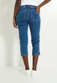 Jeans capri de mezclilla azul con bolsillos traseros, corte recto y dobladillos enrollados. Combinados con zapatillas blancas. Textura suave y de peso medio.