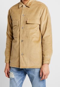 Sandfärgad kordjacka med knappstängning, två bröstfickor och lätt textur från ribbat tyg. Bärs över slitna jeans.