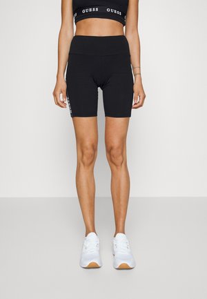 Kvinde iført sorte cykelshorts med høj talje med hvidt logo på låret og en sort sports-bh med hvidt logo-detalje, parret med hvide sneakers.