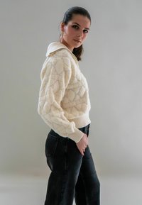 Crème-kleurige gebreide trui met een textuur van ruitpatronen en een contrasterende kraag, gecombineerd met hoogsluitende zwarte jeans.