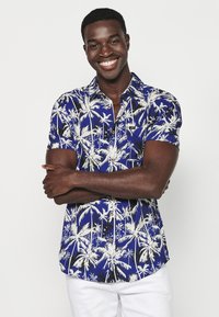 Camicia a maniche corte di colore blu con pattern di palme bianche, design con bottoni e vestibilità rilassata. Il tessuto appare leggero e morbido.