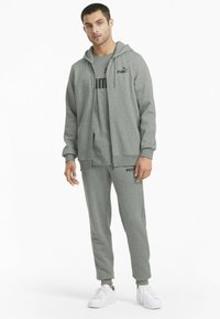 Felpa grigia con zipper e tasche, pantaloni felpa grigi abbinati, entrambi con loghi neri. Completa il look con sneakers bianche.