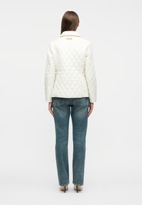 Γυναίκα που στέκεται με την πλάτη προς την κάμερα, φοράει λευκό ραμμένο μπουφάν, μπλε τζιν και μπεζ παπούτσια με ψηλό τακούνι σε λευκό φόντο.